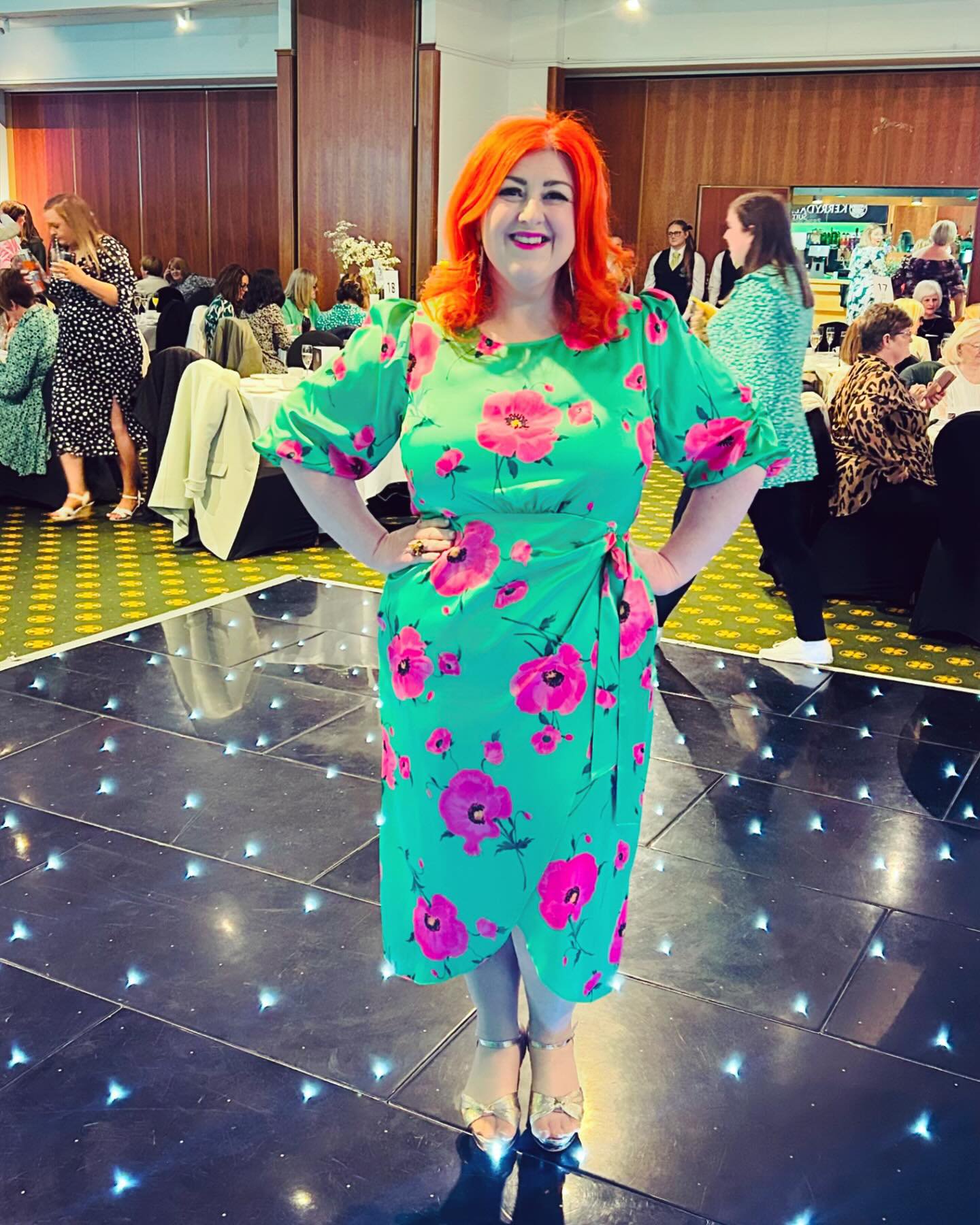 Michelle McManus smiling with bright red hair and wearing a green dress with pink floral patterns